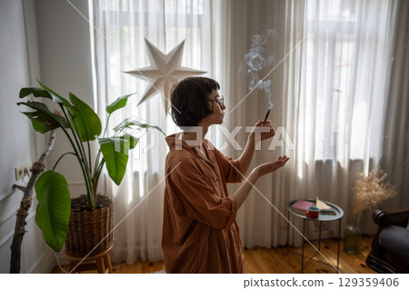 Calm woman lighting palo santo stick, fill home with aromatic smoke to create cozy atmosphere 129359406