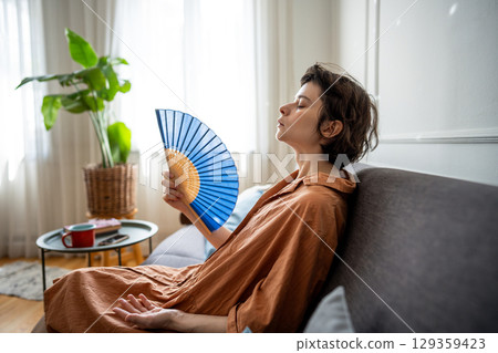 Exhausted sweaty woman fanning herself sitting on sofa suffering from summer heat without AC 129359423