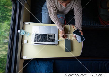 Woman plugging phone and laptop into power bank, charging batteries, sitting in train compartment. 129359439