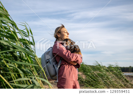 Cheerful woman holds cat in arms enjoying sunshine by riverside tall grass. Pet companionship travel 129359451