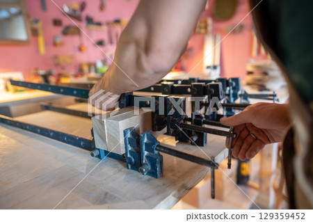Carpenter hands using clamps to press wooden beams together applying glue for board assembly for day Carpenter hands using clamps to press wooden beams together applying glue for board assembly for day 129359452