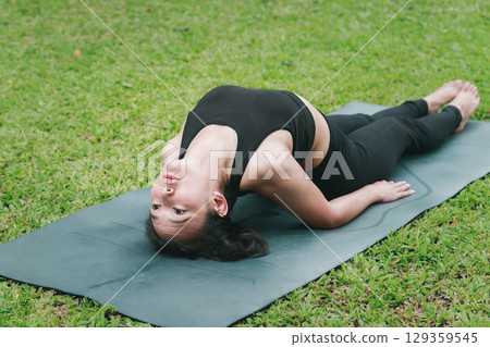 Asian woman practicing yoga in Fish pose or Matsyasana on the mat in outdoor park. Asian woman practicing yoga in Fish pose or Matsyasana on the mat in outdoor park. 129359545