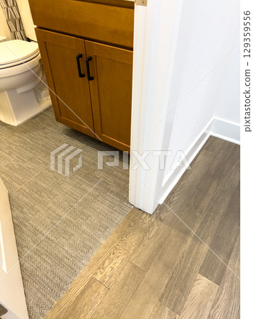A partial view of a bathroom showing a corner with a wooden vanity and white walls. The flooring is textured wood-look tile under natural lighting. A partial view of a bathroom showing a corner with a wooden vanity and white walls. The flooring is textured wood-look tile under natural lighting. 129359556