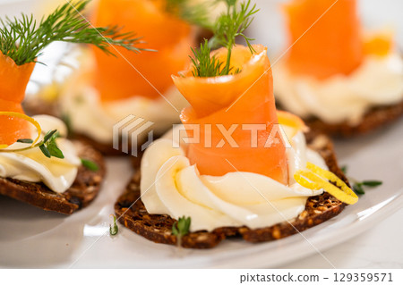A gourmet smoked salmon canape elegantly arranged on a crispy cracker, topped with a swirl of smooth cream cheese, fresh thyme, and a drizzle of olive oil. The vibrant orange salmon contrasts 129359571
