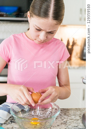 A girl stands at a kitchen counter preparing to mix ingredients in a stand mixer. She wears a pink shirt and the background includes a stove, oven, and other kitchen tools. A girl stands at a kitchen counter preparing to mix ingredients in a stand mixer. She wears a pink shirt and the background includes a stove, oven, and other kitchen tools. 129359580