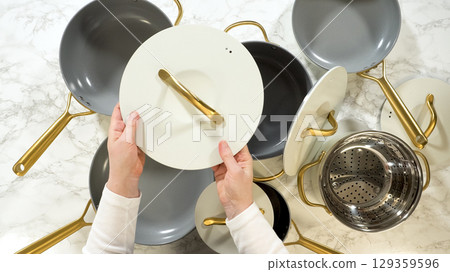 A hand holds a white pot lid with a gold handle above a gray pot. The modern cookware set is arranged on a marble countertop and includes a matching collection of pans and accessories. A hand holds a white pot lid with a gold handle above a gray pot. The modern cookware set is arranged on a marble countertop and includes a matching collection of pans and accessories. 129359596