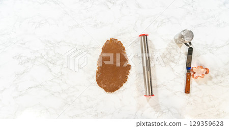 Overhead view of the dough rolling process for Spiced Gingerbread Cookies, showing hands rolling the dough on a marble surface with tools nearby. 129359628