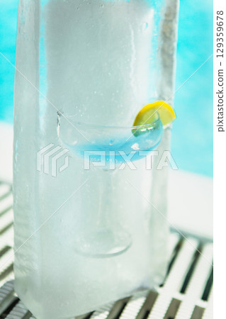 Blue cocktail in coupe glass with lemon garnish embedded in tall block of ice by pool 129359678