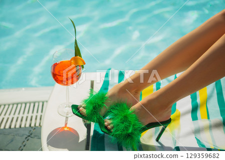 Green feathered heels and orange cocktail by swimming pool on striped towel 129359682