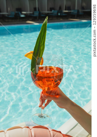 Glass of orange cocktail with leaf garnish held by poolside Glass of orange cocktail with leaf garnish held by poolside 129359698
