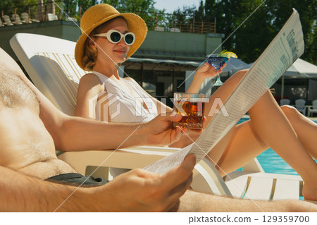 Woman in sun hat relaxing by pool with cocktail and smiling in sunlight 129359700