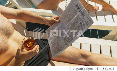 Man reading newspaper and holding drink while sunbathing by pool 129359701