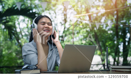 Asian woman wearing headphones listening to music on her laptop. Relaxing time. Lazy worker wasting time at workplace in co-working. 129359708