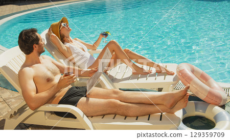 Man and woman relaxing on poolside loungers with drinks under bright summer sun 129359709
