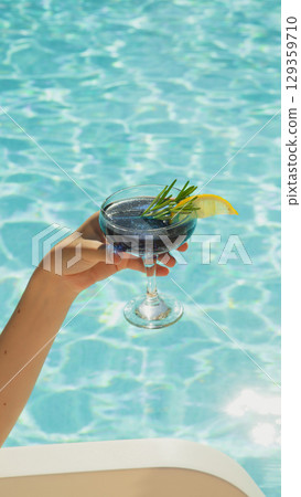 Hand holding blue cocktail with lemon and rosemary by clear swimming pool 129359710