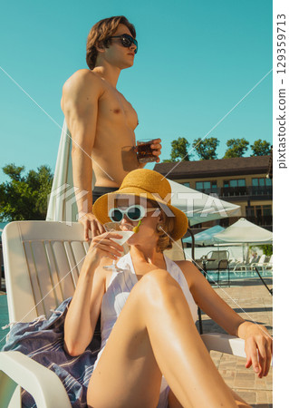 Stylish couple enjoying summer drinks by pool under bright sun 129359713