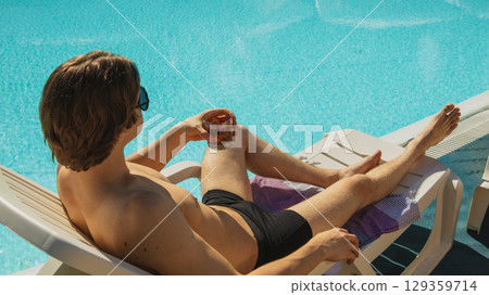 Man in black swim trunks lounging with drink by pool 129359714