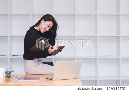Asian businesswoman sitting on the edge of desk and working in modern office. 129359762