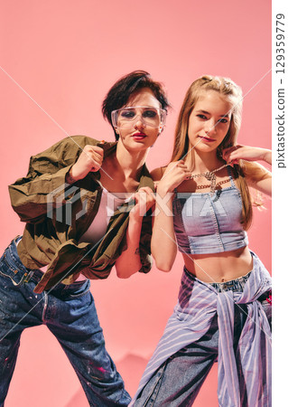 Two fashionable women in denim and casual tops posing with chains on pink background 129359779