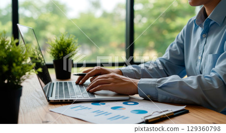 Professional businessman working on his laptop, analyzing financial data and charts in a modern, sunlit office 129360798