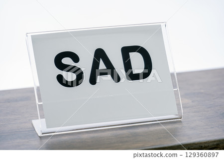 Clear acrylic sign with word SAD in bold black letters is displayed on wooden counter, conveying somber mood 129360897