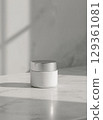 A white cosmetic container with a silver cap sits on a marble surface, with soft shadows creating depth and a serene mood. 129361081