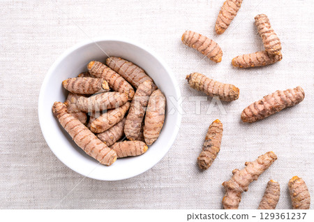 Fresh turmeric rhizomes, in a white bowl, on linen. Underground stems of the turmeric plant Curcuma longa, with bright orange flesh. Used fresh, or dried and ground as turmeric powder. Food photo. 129361237