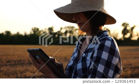 Female agronomist in straw hat monitoring harvest on barley field at sunset. Adult farmer using digital tablet on wheat meadow at dusk. Beautiful scenic landscape. Concept of agricultural business Female agronomist in straw hat monitoring harvest on barley field at sunset. Adult farmer using digital tablet on wheat meadow at dusk. Beautiful scenic landscape. Concept of agricultural business 129361453