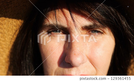 Face of adult woman in straw hat illuminating by bright rays of sun. Tired blue eyes of female blinking and looking into camera outdoor. Upset facial expression of lady. Close up Slow motion 129361455