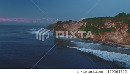 Soft pink and purple hues paint the sky as the sun sets over the dramatic cliffs of Pantai Dreamland Beach in Bali, Indonesia, with waves crashing against the rocky shore 129361835