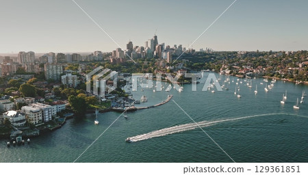 Australia, Sydney: Motorboat cruising Sydney Harbour, leaving white trail. Lavender Bay with sailboats yachts against the modern urban architecture city skyline in sunny day. Aerial view drone 129361851
