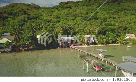 Tranquil lagoon waters lapping wooden pier lead to cozy beachfront houses nestled among lush tropical vegetation and a majestic verdant hillside. Tahiti tropical island, French Polynesia. Drone flight 129361863