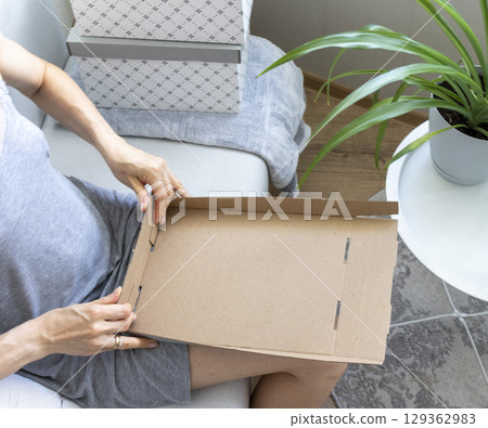 Woman sitting in the couch in cozy room, assembling carton box. Indoors 129362983