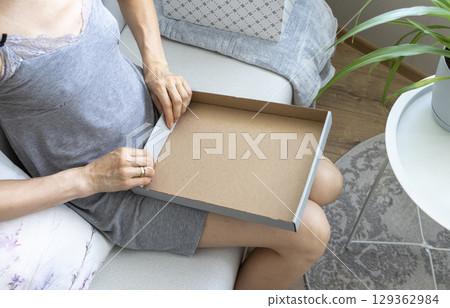 Woman sitting in the couch in cozy room, assembling carton box. Indoors 129362984