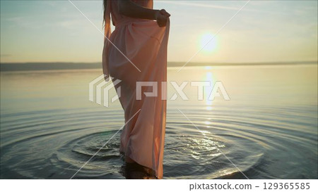 Young woman in white dress walking alone on the beach in the sunset. girl in a dress stands in the water. 129365585