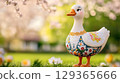 Goose figurine with embroidered dress, stands in garden setting with flowers background, copy space 129365666