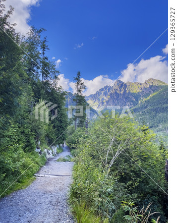 Landscape in the foothills of the Tatras. The peaks of mountains against the blue sky and clouds are visible from the mountain path. Landscape in the foothills of the Tatras. The peaks of mountains against the blue sky and clouds are visible from the mountain path. 129365727