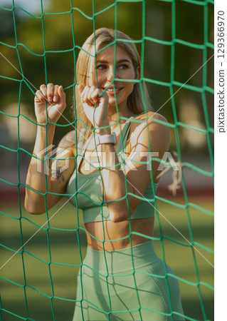 young woman in activewear smiles while leaning against a green sports net She enjoys an outdoor workout under clear skies, exuding positivity and energy young woman in activewear smiles while leaning against a green sports net She enjoys an outdoor workout under clear skies, exuding positivity and energy 129366970