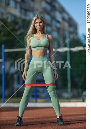 sporty girl engages in strength training exercises outdoors, utilizing a red elastic band for resistance The scene captures her focused expression as she works out in an urban area 129366989