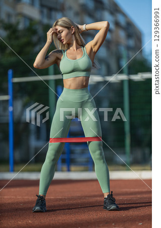 sporty girl in form fitting green workout attire uses red elastic band for resistance training at outdoor fitness area setting features workout equipment and greenery 129366991