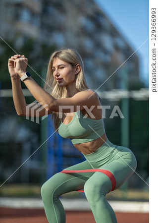 Sporty girl performs squat exercise outdoors on training court is dressed in green activewear and uses red elastic band to enhance her workout 129366993