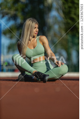 A fitness enthusiast sits cross legged on a track at a sports facility, enjoying the sunny weather while engaging in stretching The scene captures her athletic attire and relaxed posture A fitness enthusiast sits cross legged on a track at a sports facility, enjoying the sunny weather while engaging in stretching The scene captures her athletic attire and relaxed posture 129367003