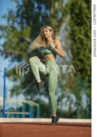 sporty girl jogs in place on a bright sunny day at a local park She wears a green fitness outfit and is focused on maintaining her workout, enjoying the fresh air and scenery 129367008