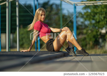 fitness enthusiast sits on a wooden platform at an outdoor gym, enjoying the warm light of late afternoon She wears a fitted top and shorts, showcasing her tattoos and athletic physique fitness enthusiast sits on a wooden platform at an outdoor gym, enjoying the warm light of late afternoon She wears a fitted top and shorts, showcasing her tattoos and athletic physique 129367023