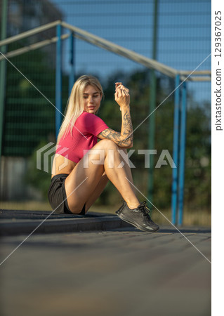 young woman sits on a track in a sports field, wearing a stylish athletic outfit, displaying tattoos, and enjoying a moment of relaxation in the sun 129367025