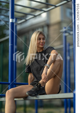 person with long blonde hair sits casually on outdoor gym equipment, stretching one leg while enjoying the warm glow of golden hour in a modern urban setting 129367038