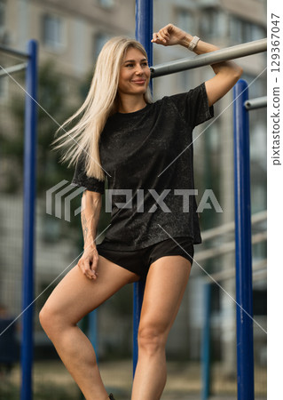 woman with long blonde hair relaxes at an outdoor gym, leaning against exercise bars She wears a black shirt and shorts, enjoying a sunny afternoon in an urban setting woman with long blonde hair relaxes at an outdoor gym, leaning against exercise bars She wears a black shirt and shorts, enjoying a sunny afternoon in an urban setting 129367047
