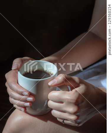 Close up of Comforting cozy female hands holding a coffee glass with the morning sunlight that shines through. 129368154