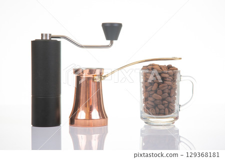 A black manual coffee grinder, a copper cezve with a gold handle, and a glass mug A black manual coffee grinder, a copper cezve with a gold handle, and a glass mug 129368181