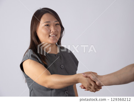 A woman with dark hair and a gray top is shaking hands with another person, whose arm and hand A woman with dark hair and a gray top is shaking hands with another person, whose arm and hand 129368212